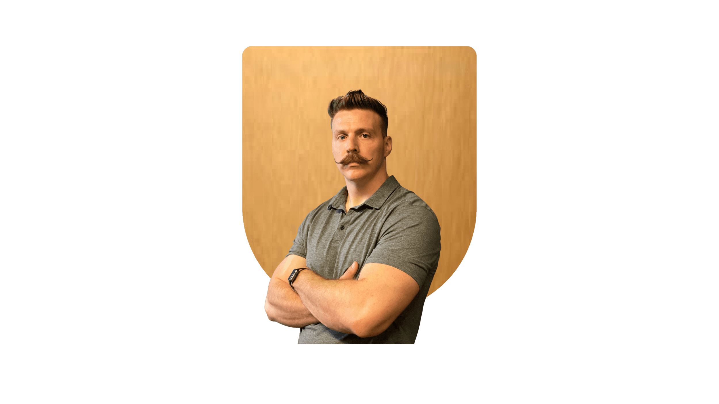 A young man with styled hair and a curled moustache, wearing a grey polo shirt, poses with his arms crossed against a wooden background.