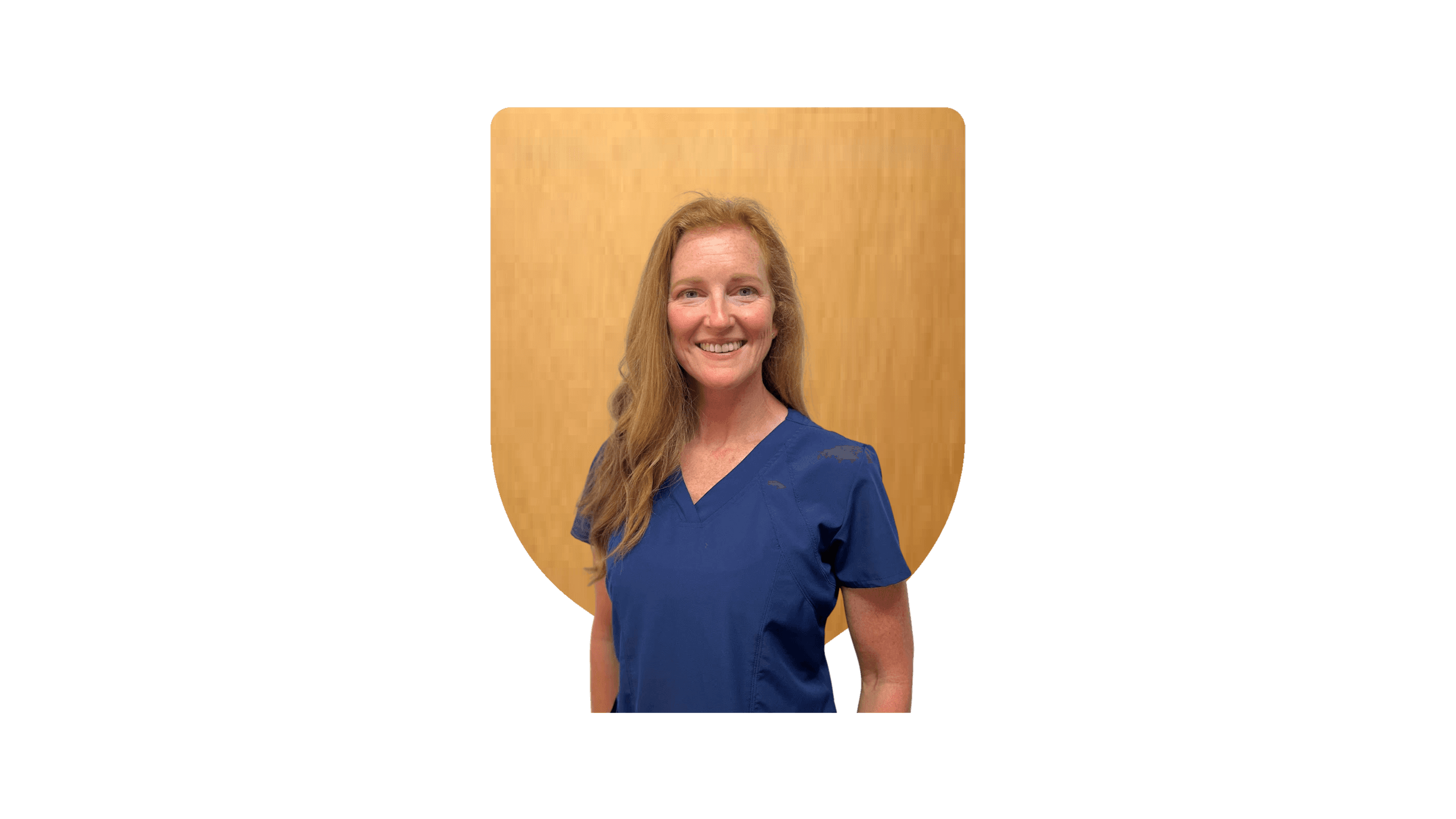 Portrait of a smiling woman with long, wavy red hair wearing a navy medical scrub top, standing in front of a wooden background.